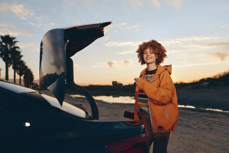Woman lifestyle scene on road trip at sunset, young woman in orange jacket stands by car with open hood and backpack, curly hair smiling outdoors, golden sky travel mood naturally