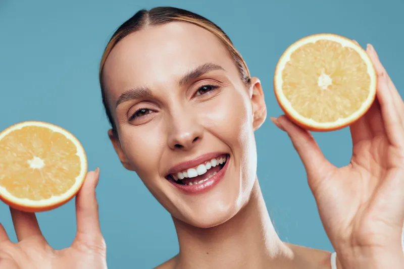 Smiling woman holding two halves of fresh grapefruit near her face isolated on blue background with healthy skin and joyful expression closeup portrait.