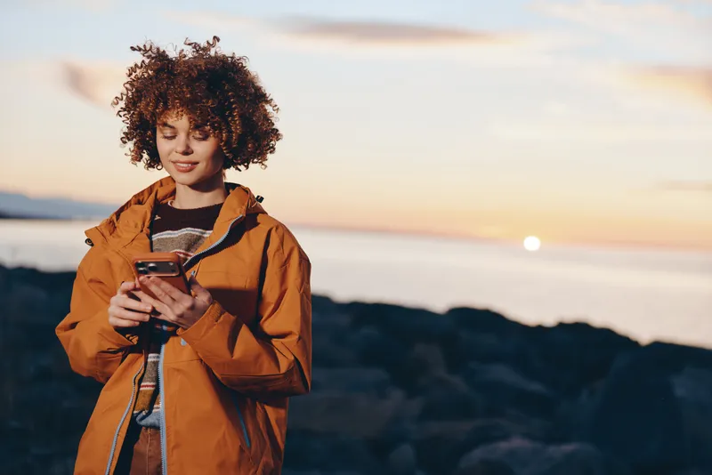 Woman lifestyle portrait on beach at sunset, curly hair and orange jacket, holding camera outdoors, travel atmosphere by the sea coast, dusk sky, relaxed smile, lifestyle concept