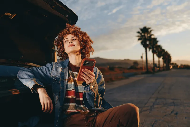 Woman lifestyle, using smartphone by open car door, curly hair, denim jacket, warm sunset, outdoor road with palm trees, relaxed travel moment, casual street style