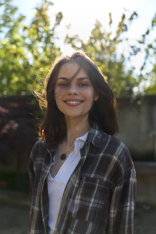 Young woman with long dark hair smiles softly outdoors in a plaid shirt, sunlight on her face, capturing a casual, relaxed everyday moment and natural confidence