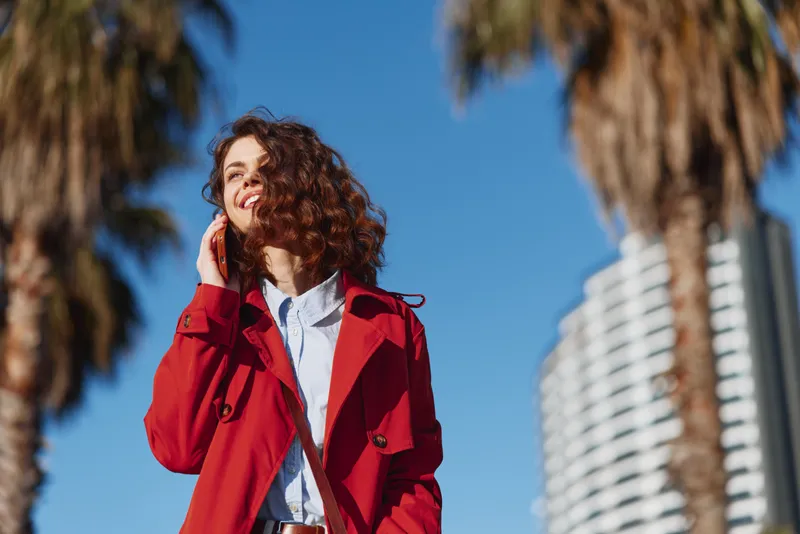 A stylish woman in a red coat stands outdoors under a bright blue sky, smiling softly. Modern architecture and palm trees frame her, conveying bold street fashion and confidence.