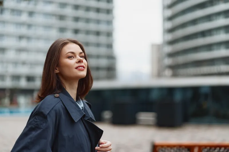 A poised young woman in a dark trench coat gazes upward in a modern urban plaza, conveying confidence, contemplation and curiosity amid glass buildings.