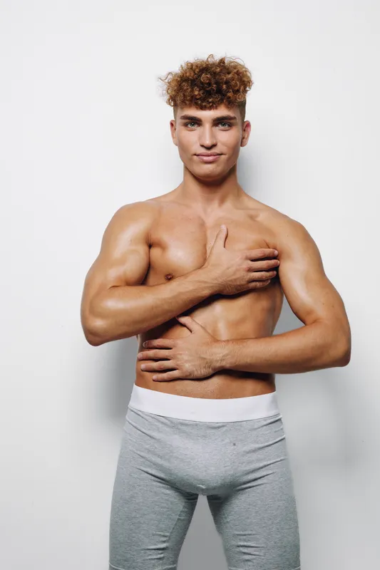 Young athletic man posing shirtless with muscular body wearing gray pants while standing against studio wall with confident expression and healthy lifestyle vibes.