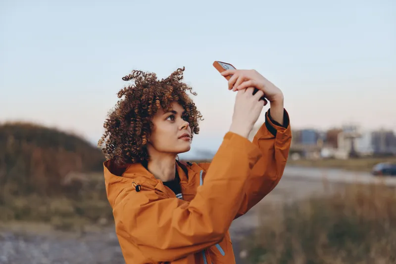 Woman lifestyle outdoors portrait, curly hair, orange jacket, looking into distance, one hand shading eyes, calm thoughtful expression in warm sunset light, landscape field behind