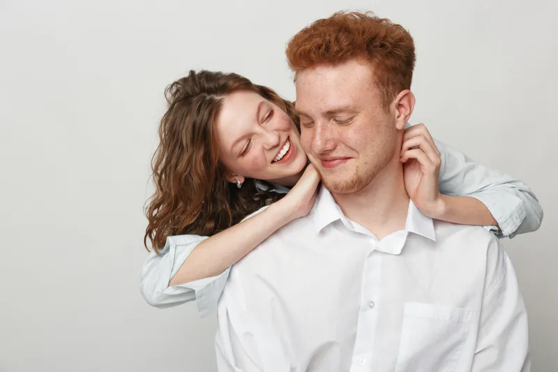 Happy young couple in casual white shirts share a tender moment, woman embraces man from behind with eyes closed. Concept of love, affection, intimacy, and relationship happiness.