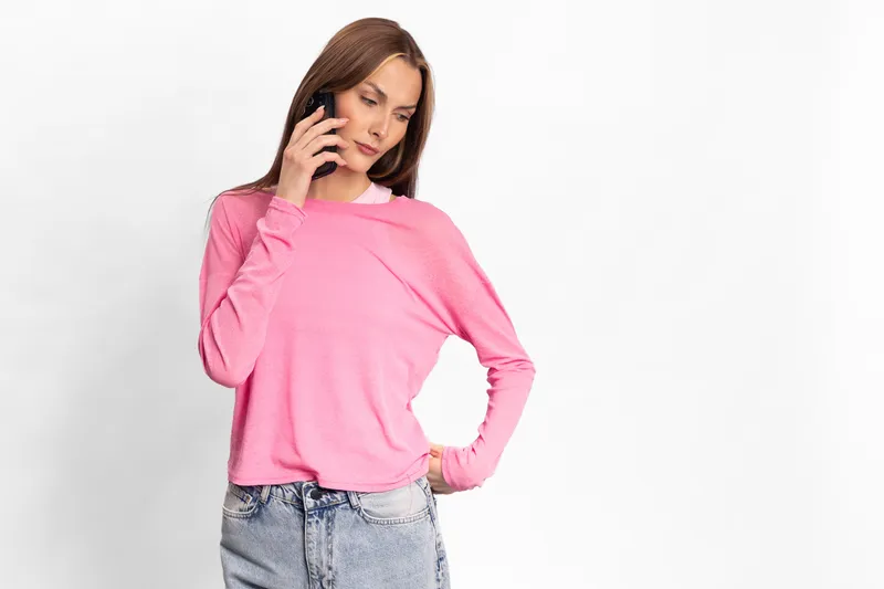 Young woman in pink sweater and blue jeans talking on smartphone with serious expression, isolated on white background. Concept of communication, technology, and casual lifestyle.