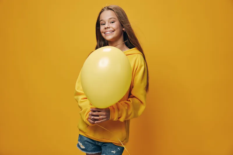 Smiling girl with long hair wearing yellow hoodie and denim shorts holding yellow balloon on yellow background for celebration and happiness.