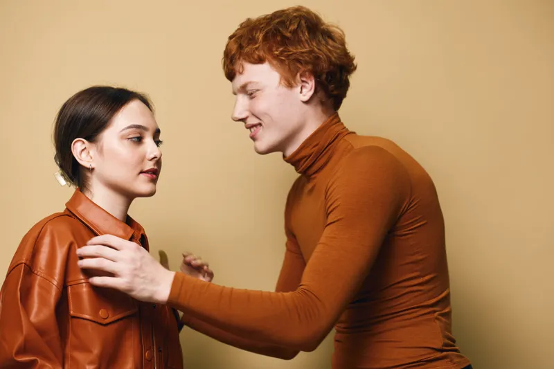 Young man and woman interacting indoors with focused expressions, wearing casual autumn outfits, showcasing human connection and warm tones in soft lighting.