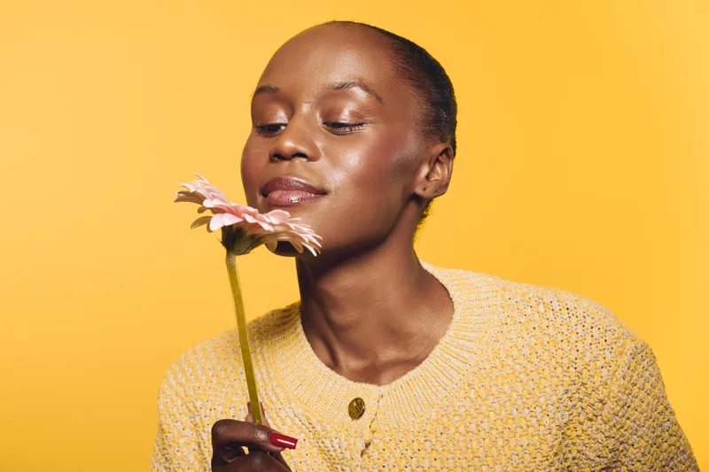 Portrait of a dark skinned African American woman with closed eyes holding a pink flower near her face. She wears a yellow knitted sweater against a yellow background. Concept of serenity and beauty.