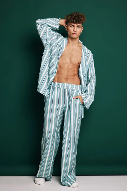 Young man posing wearing striped sleepwear with open shirt, standing with serious expression on dark green background showing bare chest and pants with pockets.