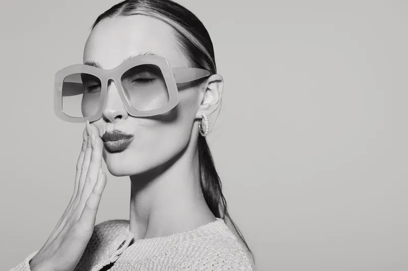 Stylish slim Caucasian woman with pale skin wearing large sunglasses and light sweater blows a kiss with closed eyes in studio. Concept of fashion, beauty, and elegance.