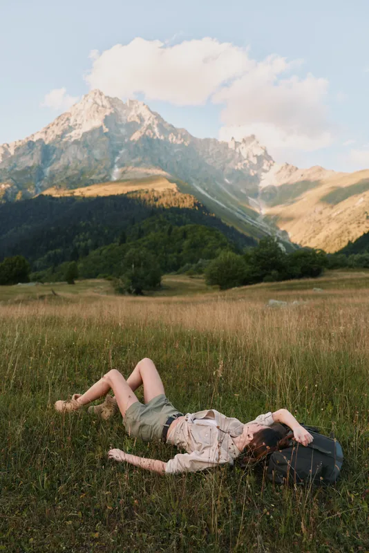 A woman in casual clothes lies on grass in a mountain meadow resting with a backpack nearby under a clear sky. The scene shows outdoor lifestyle and relaxation in nature.