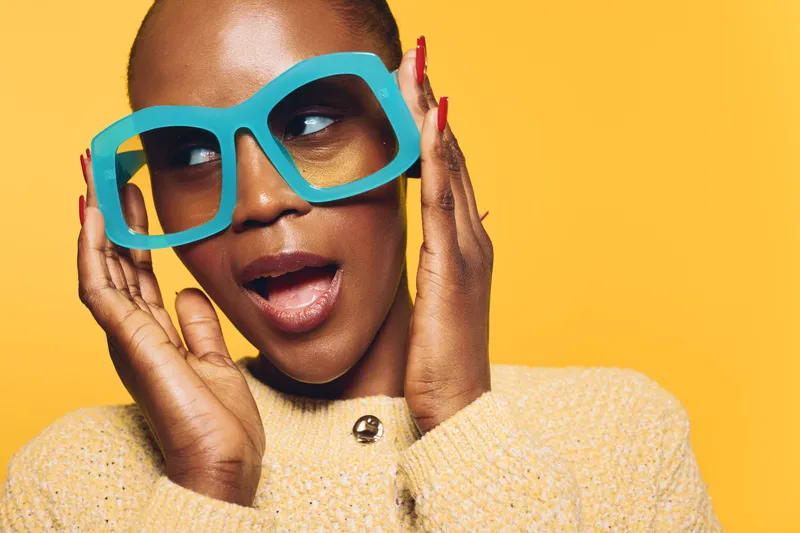 Close-up of a surprised dark skinned African woman wearing oversized blue glasses and a beige sweater on yellow background. Concept for fashion, expression, and colorful accessories.