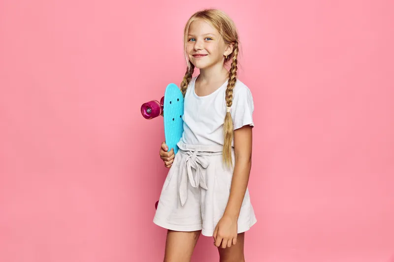 Young girl with braided hair stands holding blue skateboard on pink background smiling confidently. Casual outfit with white shirt and beige shorts.
