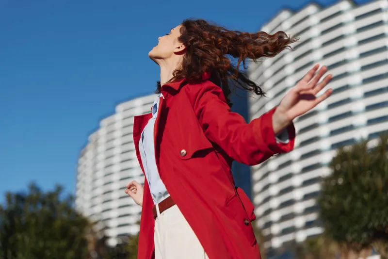 Young woman in a red coat lifts her face to the sun, arms outstretched, wind in her hair, with a bright city backdrop of tall buildings and blue sky behind her.
