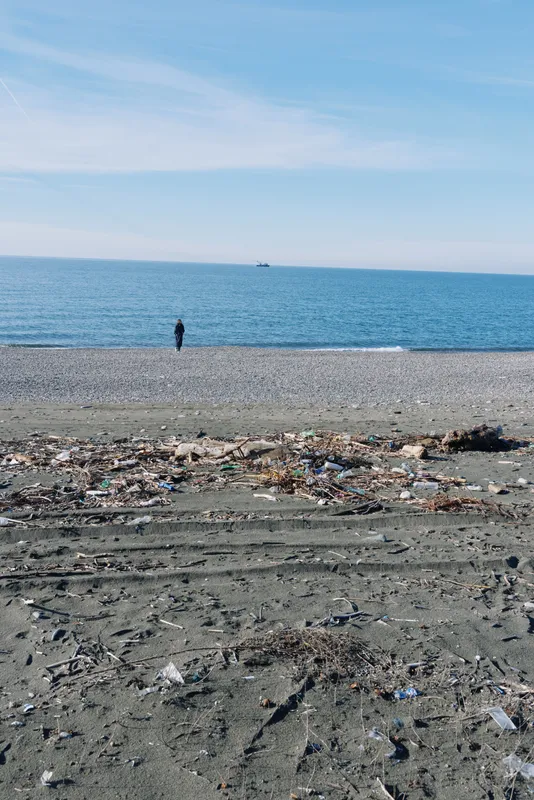 Beach scene with a lone figure walking along a pebble shore, litter scattered across the sand, exposing pollution and discarded debris by the calm ocean and water