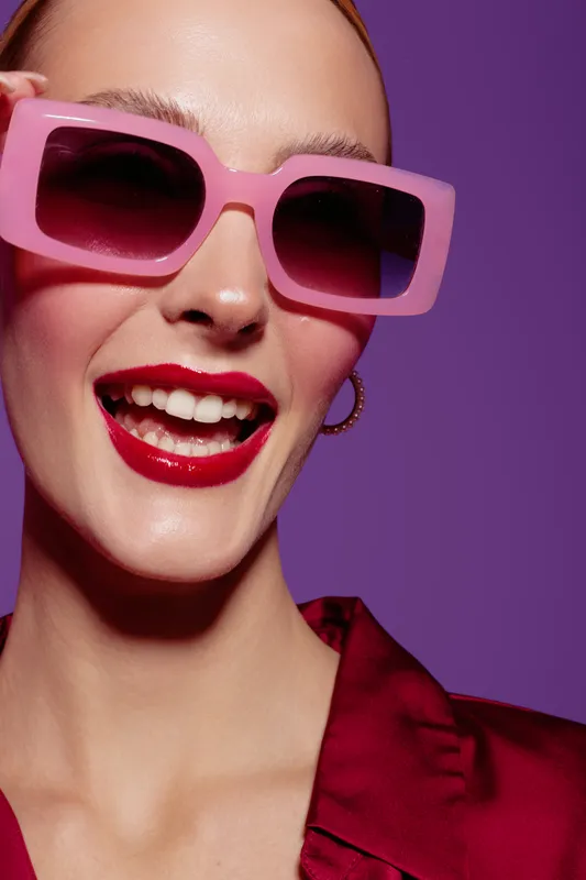 Close-up of a happy woman wearing large pink sunglasses and red lipstick smiling against a purple background. Concept of fashion, beauty, summer style, and joyful mood.