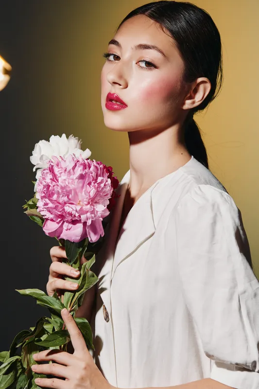 Young woman with flawless skin and bold lipstick holds pink peony flower, posing elegantly in bright blouse against soft beige background. Beauty fashion concept.