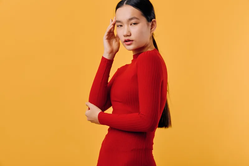 Young woman in red dress posing with hand near face against orange background. Asian female model with long hair looking at camera in studio with casual confident expression for fashion.