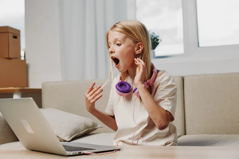 Surprised blonde girl with purple headphones around neck reacts to laptop screen while sitting on beige sofa in bright modern room with natural light and cardboard boxes in background.