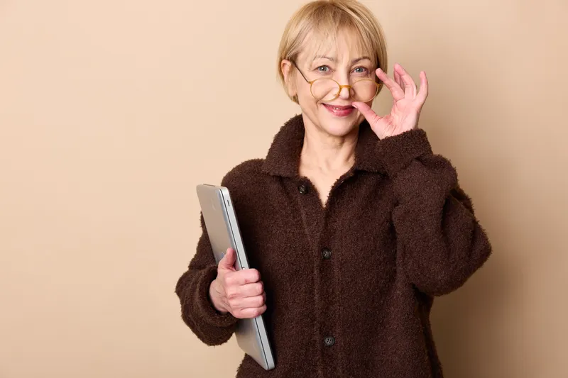 Elderly woman with glasses smiles and waves at camera while holding a tablet, studio portrait on beige background, friendly technology communication senior