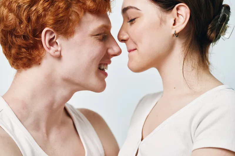 Young man and woman facing each other closely with eyes closed, smiling and enjoying intimate moment in studio with white background and casual clothing.
