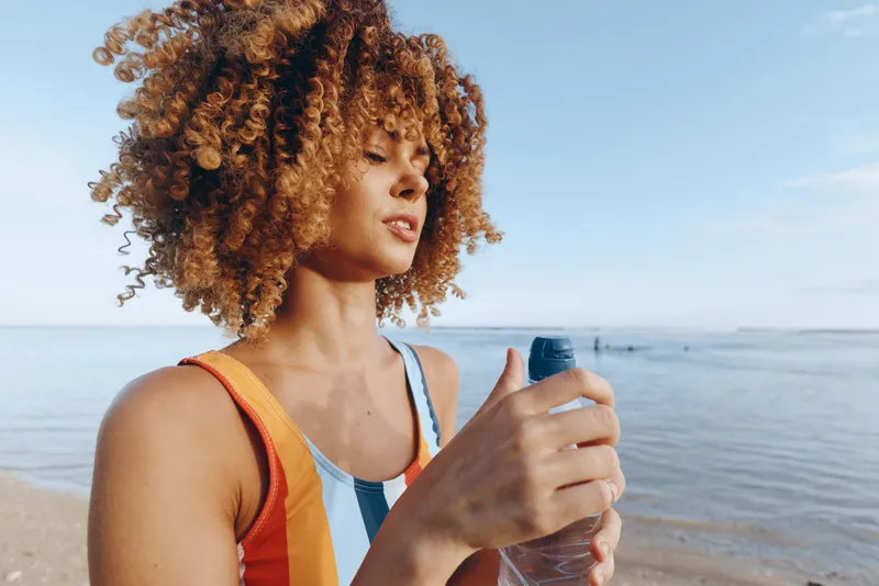 Young african american woman with curly hair in colorful swimsuit opens a bottle of water while standing on a sandy beach under clear blue sky near calm sea.