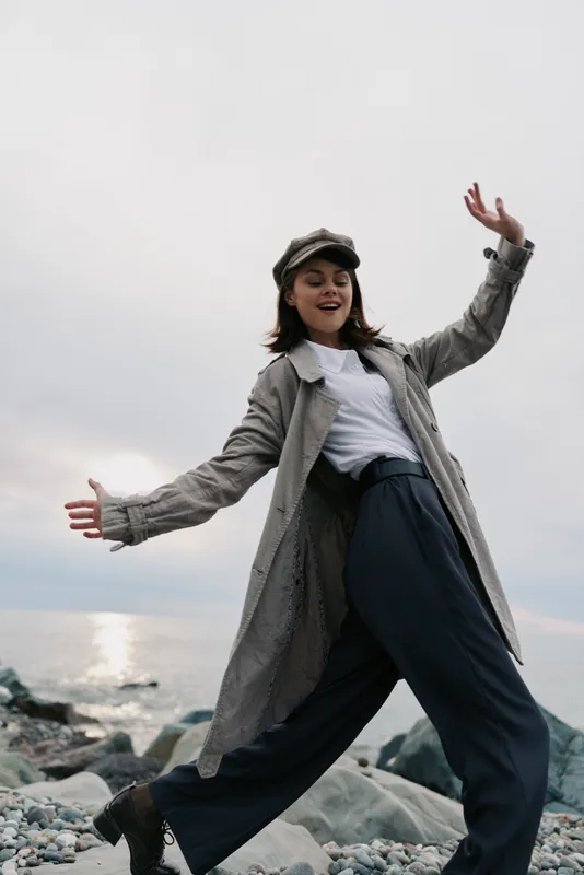 Young woman wearing grey coat and hat enjoys windy day on rocky beach. She poses with arms outstretched near water under cloudy sky. Concept of freedom, fashion, and outdoor lifestyle.