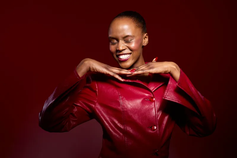 Smiling black woman with dark skin in red leather jacket poses with hands under chin on dark red background. Concept of fashion, beauty, confidence, and joyful mood.