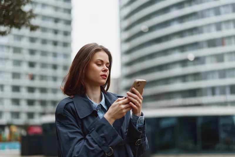 A focused young woman in a dark coat uses a smartphone outdoors in an urban environment with modern buildings blurred in the background, representing technology and city life.