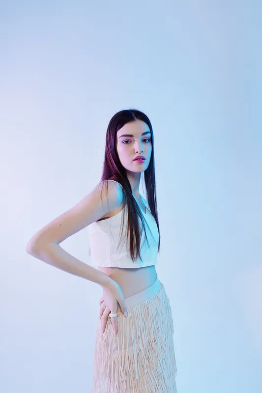 Young woman beauty fashion model poses in a white crop top and fringed skirt. Studio shot with soft lighting on a light background. Concept of style and elegance.