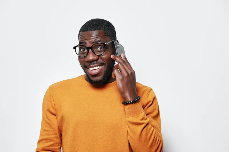 African American man with dark skin wearing glasses and orange sweater talking on smartphone smiling isolated on white background with positive expression and communication.