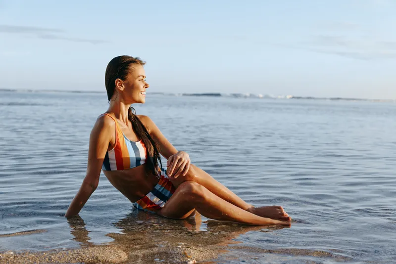 Woman at beach ocean swimsuit sitting in shallow water under sunlight, calm relaxation in summer near the shoreline, gentle waves and distant horizon create a peaceful outdoor lifestyle.