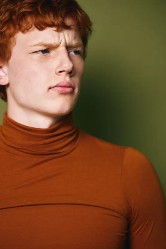 Young man with red hair in orange turtleneck looking aside with serious expression on green background for portrait and fashion concept.