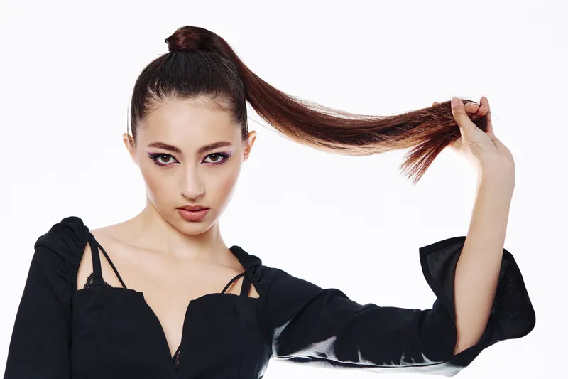 Fashion model with sleek high ponytail holds hair with one hand wearing a black top on a white background. Beauty and creative concept with striking makeup and intense gaze.