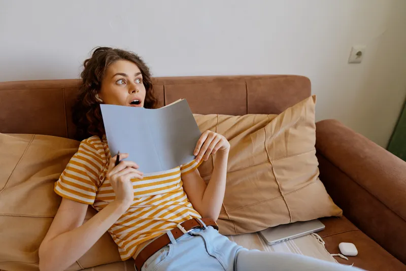 Woman lifestyle indoors, reading a notebook on a couch, striped top, surprised expression, cozy living room, sunny window glow, soft textures, relaxed posture, warm colors, home atmosphere