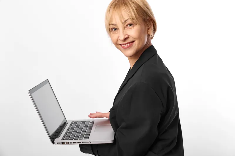 Smiling older woman in black business suit with laptop on white background, standing in front view, calm confident expression, workplace technology concept, isolated portrait