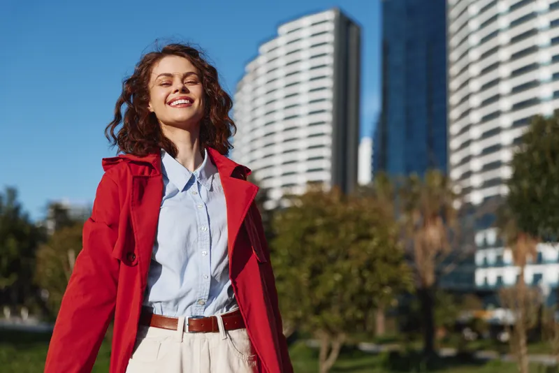 Young woman in a red coat stands outdoors in an urban park, smiling with sunlight on her face as modern buildings rise behind her.