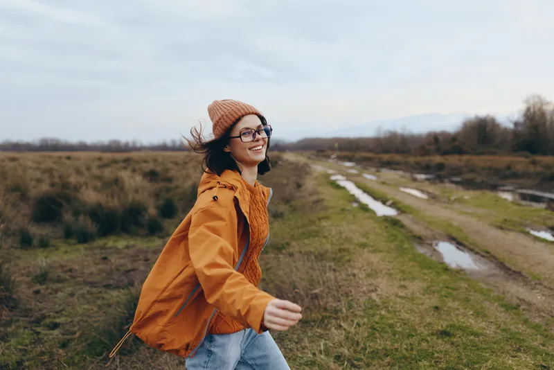Smiling woman in orange jacket and knit beanie walks along a grassy country path, winter landscape, outdoors lifestyle, natural light, casual street fashion, calm mood, fresh air day trip