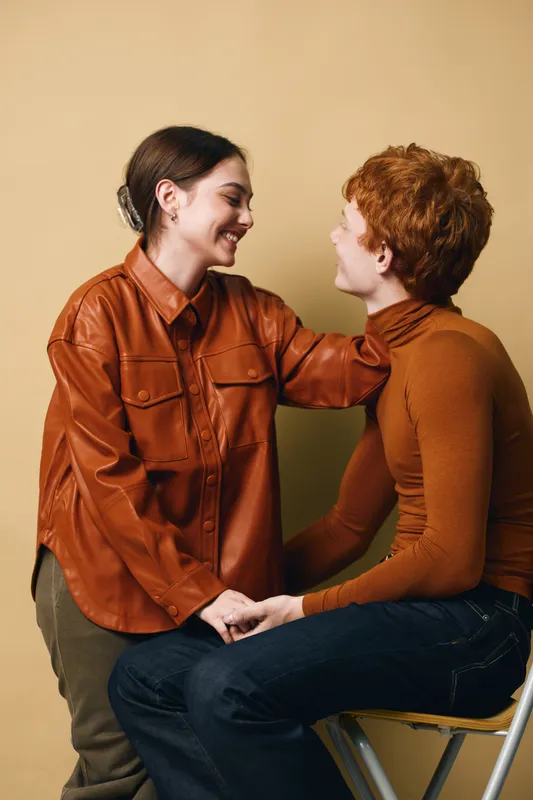 Couple sitting together indoors, smiling and holding hands while looking at each other. Woman with brown leather shirt and man with orange sweater sharing affection and warmth in cozy room.