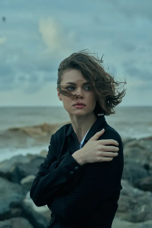 Woman in dark attire by a windy shoreline, hair whipping, moody coastal atmosphere, conveying resilience, fashion mood, and cinematic emotion against rough water.