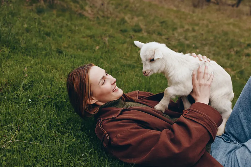 A relaxed woman rests on the grass, cuddling a playful goat kid in a sunny outdoor moment. A candid lifestyle scene that highlights bonding with animals and simple joy in a rural setting.