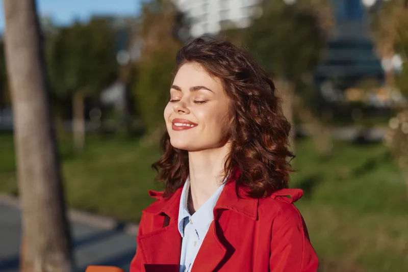 A smiling woman in a red coat stands outdoors in bright sun, eyes closed, enjoying warm weather. Concept: confidence, happiness, and carefree mood.