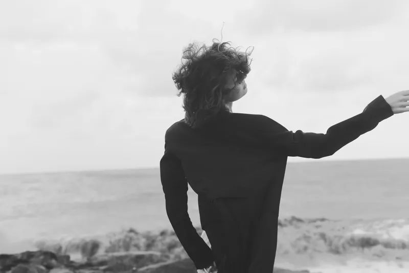Woman on a windswept beach extends her arm, creating a dramatic silhouette against the ocean horizon, conveying freedom, solitude, and contemplative coastal beauty in monochrome tones under soft