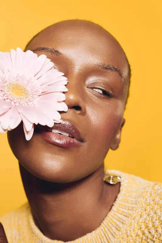 Close-up of an African woman with dark skin holding a pink flower near her face, wearing a yellow sweater. The bright yellow background creates a warm, vibrant mood of natural beauty and wellness.