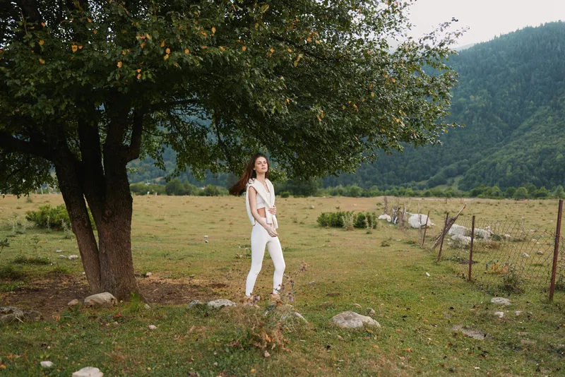 Young woman in a white jumpsuit stands beside a large tree in a sunlit field with distant mountains, conveying calm, freedom, and a relaxed lifestyle.