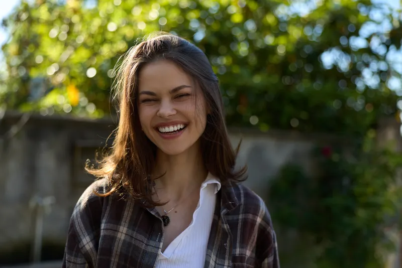 Joyful outdoor lifestyle portrait of a woman smiling in sunlight, wearing casual plaid shirt, relaxed pose, natural garden backdrop, candid moment of everyday happiness, and optimism