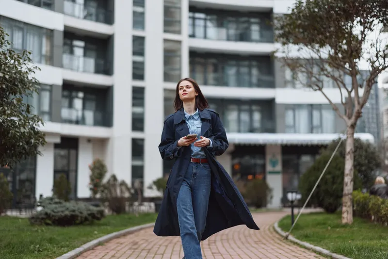 Young woman in a denim coat and jeans walks through a modern residential courtyard, exuding confident street style in natural daylight, a candid everyday moment.
