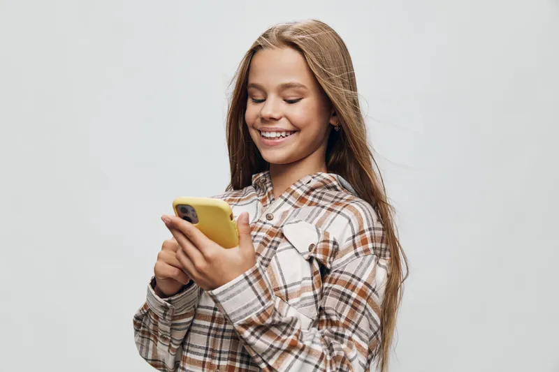 Teenage girl smiling and looking at smartphone while wearing plaid shirt and backpack on gray background. Casual style, technology and youth with positive expression.